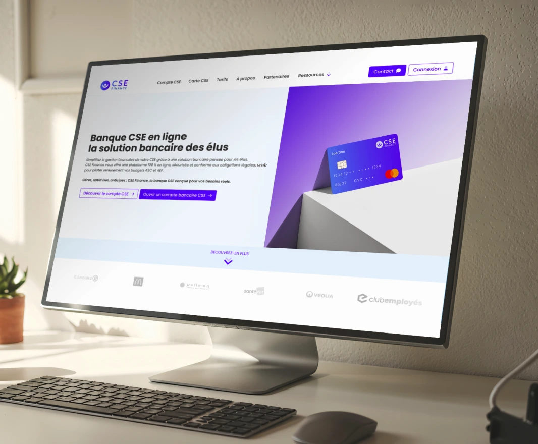 Desktop monitor on a desk displaying a French online banking homepage with a purple card image and navigation bar, marketing text visible on the screen